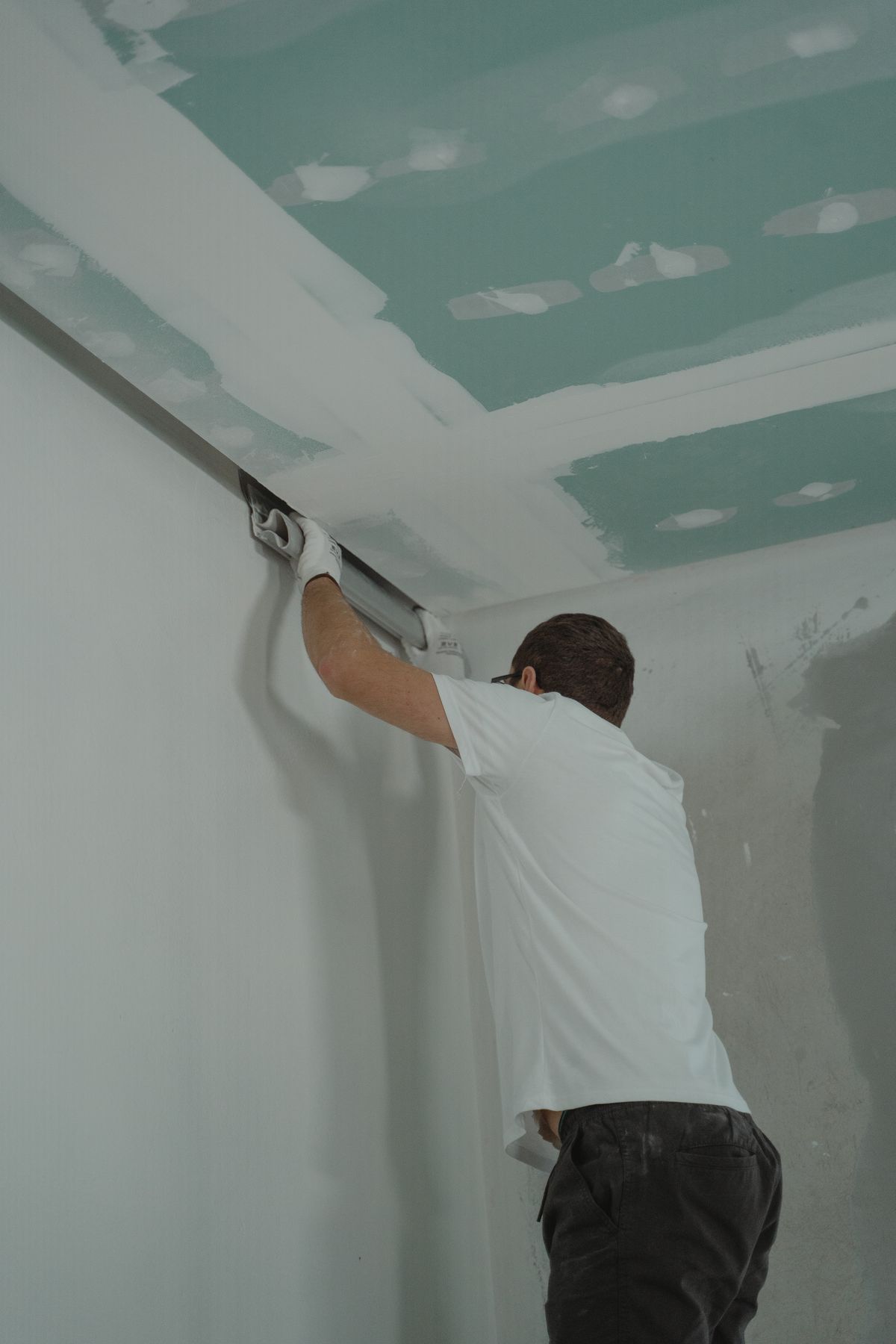 Worker pulling a wide finishing knife along the joint where a taped ceiling meets the wall