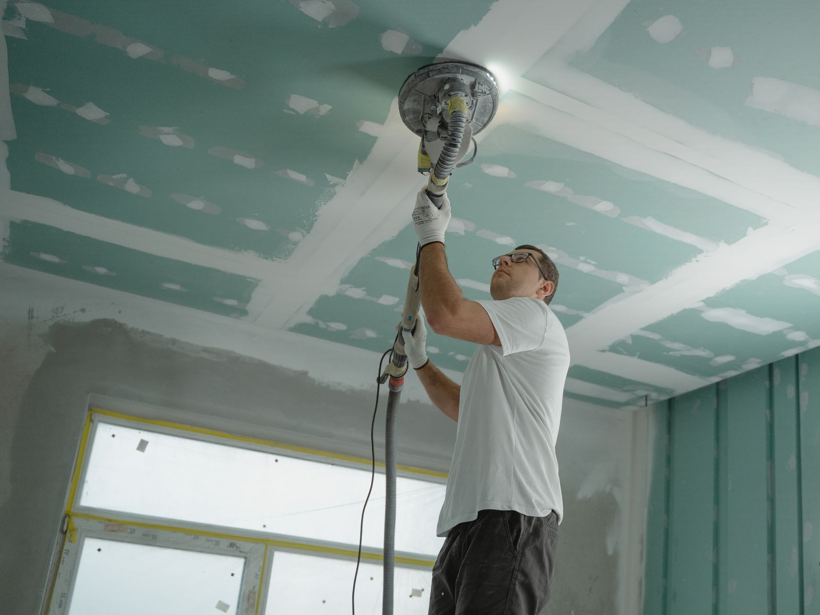 Worker using a dust-extracting ceiling sander to smooth a taped and mudded drywall ceiling